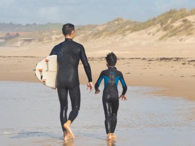 Father and son in wetsuits walk along Portugal beach, surfboard in hand, creating a memorable surfing adventure.