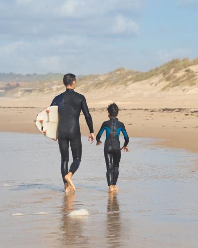 Father and son in wetsuits walk along Portugal beach, surfboard in hand, creating a memorable surfing adventure.