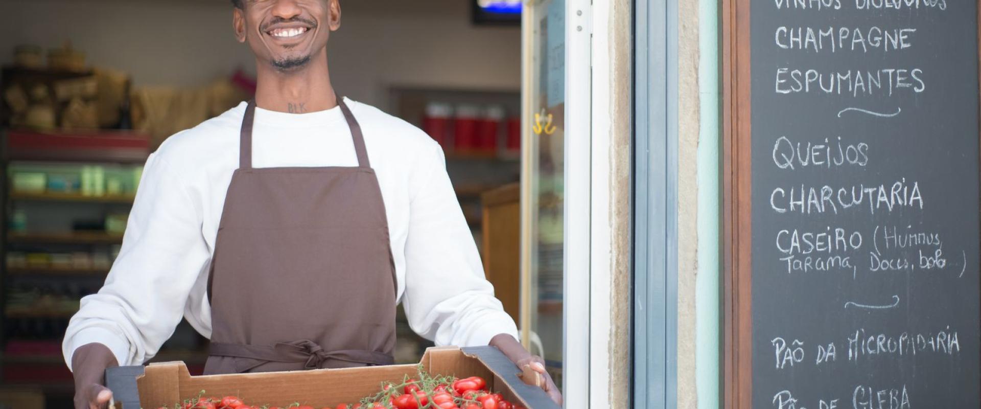 Smiling vendor standing outside a Portuguese store holding a box of ripe tomatoes. Perfect for grocery or food themes.