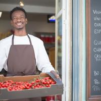 Smiling vendor standing outside a Portuguese store holding a box of ripe tomatoes. Perfect for grocery or food themes.