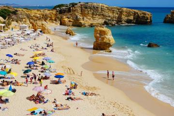 A crowded sandy beach with numerous colorful umbrellas and sunbathers, backed by golden limestone cliffs and rock formations under a clear blue sky, with turquoise water and gentle waves.