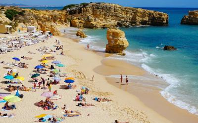 A crowded sandy beach with numerous colorful umbrellas and sunbathers, backed by golden limestone cliffs and rock formations under a clear blue sky, with turquoise water and gentle waves.