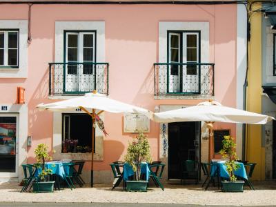 A street-level view of a pink building facade with two white-framed windows featuring black iron balconies, a central entrance, and an outdoor seating area with white umbrellas and blue tables on a cobblestone sidewalk.