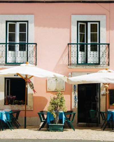 A street-level view of a pink building facade with two white-framed windows featuring black iron balconies, a central entrance, and an outdoor seating area with white umbrellas and blue tables on a cobblestone sidewalk.