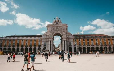 A crowd of tourists walking on a patterned stone street towards a large triumphal arch in a European city square.