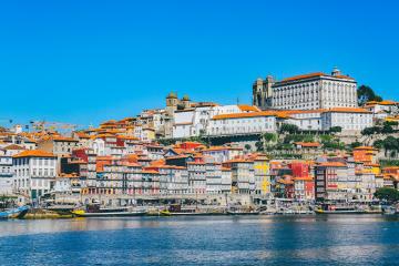 A sunlit riverside scene featuring traditional Rabelo boats on the Douro River, with colorful multi-story buildings lining the steep hillside of the Ribeira district.
