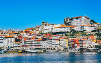 A sunlit riverside scene featuring traditional Rabelo boats on the Douro River, with colorful multi-story buildings lining the steep hillside of the Ribeira district.