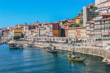 A yellow vintage tram travels down a narrow, sunlit cobblestone street lined with historic buildings featuring arched colonnades and traditional Portuguese architecture.