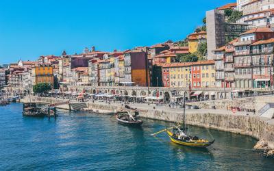 A yellow vintage tram travels down a narrow, sunlit cobblestone street lined with historic buildings featuring arched colonnades and traditional Portuguese architecture.