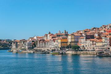 A panoramic view of the Ribeira district in Porto, Portugal, showing colorful historic buildings lining the Douro River with boats docked along the quay under a clear blue sky.