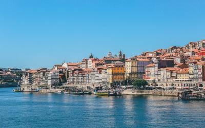 A panoramic view of the Ribeira district in Porto, Portugal, showing colorful historic buildings lining the Douro River with boats docked along the quay under a clear blue sky.