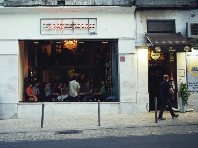 The image shows the exterior of a wine bar with a large window revealing patrons seated inside, a neon sign reading "Comida Independente" above the window, and a person walking past on the sidewalk; the adjacent business has a brown awning with "B
