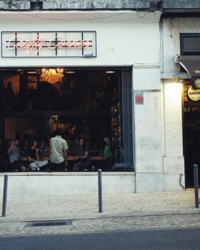The image shows the exterior of a wine bar with a large window revealing patrons seated inside, a neon sign reading "Comida Independente" above the window, and a person walking past on the sidewalk; the adjacent business has a brown awning with "B