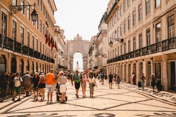 A wide, sunlit pedestrian street paved with black-and-white wave-patterned cobblestones, lined with multi-story buildings featuring balconies and flags, leading toward a large triumphal arch in the distance.