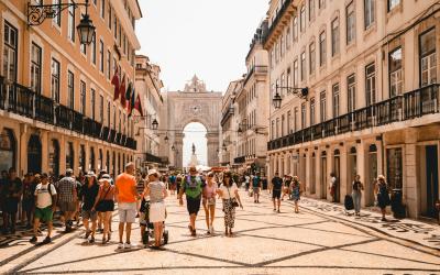 A wide, sunlit pedestrian street paved with black-and-white wave-patterned cobblestones, lined with multi-story buildings featuring balconies and flags, leading toward a large triumphal arch in the distance.