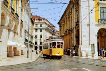 A yellow vintage tram travels down a narrow, cobblestone street lined with historic buildings featuring arched colonnades and balconies, with overhead electric wires and pedestrians visible on the sidewalks.