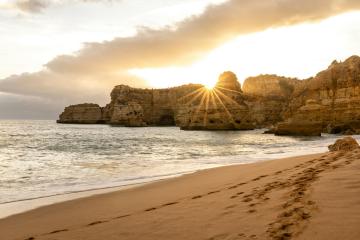 A coastal scene at sunset or sunrise featuring golden sand in the foreground, calm ocean water, and dramatic limestone cliffs with a natural arch formation silhouetted against the sun.