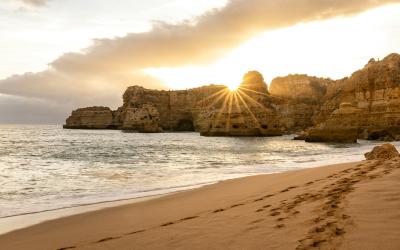 A coastal scene at sunset or sunrise featuring golden sand in the foreground, calm ocean water, and dramatic limestone cliffs with a natural arch formation silhouetted against the sun.