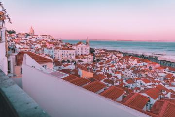 A high-angle view of densely packed white buildings with terracotta-tiled roofs overlooking the ocean at dusk, featuring a prominent church tower and a distant dome on a hill.