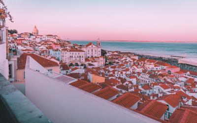A high-angle view of densely packed white buildings with terracotta-tiled roofs overlooking the ocean at dusk, featuring a prominent church tower and a distant dome on a hill.
