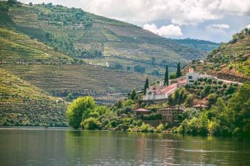 A white-walled estate with a red-tiled roof nestled on a steep, terraced hillside overlooking a wide river, with extensive vineyards climbing the slopes under a partly cloudy sky.