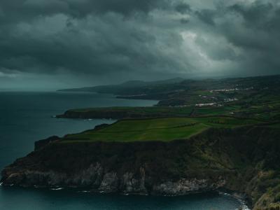 A dramatic coastal landscape featuring steep, dark cliffs dropping into the ocean, with a strip of green pastureland along the cliff edge and a cloudy, stormy sky overhead.