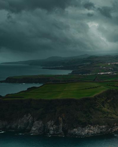 A dramatic coastal landscape featuring steep, dark cliffs dropping into the ocean, with a strip of green pastureland along the cliff edge and a cloudy, stormy sky overhead.