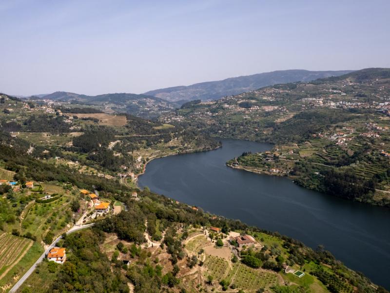 A wide-angle landscape photograph showing a large river curving through a hilly, terraced valley with scattered houses and vineyards under a clear sky.