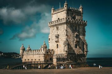 A large, ornate stone tower with crenellated battlements and a decorative armillary sphere on top, situated on a small island connected to the shore by a walkway, with people walking on the sand and water in the background.