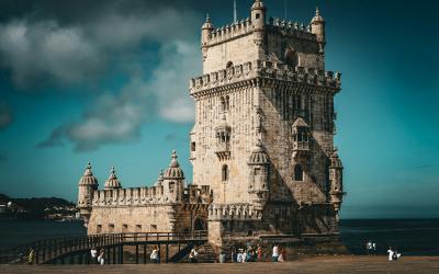 A large, ornate stone tower with crenellated battlements and a decorative armillary sphere on top, situated on a small island connected to the shore by a walkway, with people walking on the sand and water in the background.