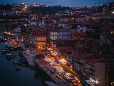 A high-angle view of the Ribeira district in Porto, Portugal, at twilight, showing the Douro River, traditional buildings, and outdoor dining areas along the quay.