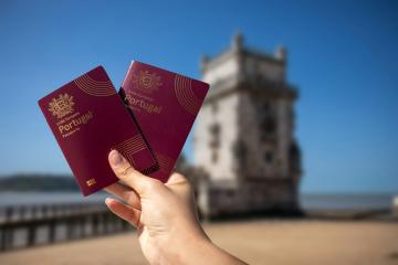 A hand holding two burgundy Portuguese passports in the foreground with the Belém Tower in Lisbon blurred in the background.