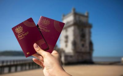 A hand holding two burgundy Portuguese passports in the foreground with the Belém Tower in Lisbon blurred in the background.