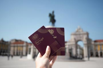 A hand holding two maroon passports with "Portugal" printed on the cover, set against a blurred background of a large plaza with a triumphal arch and a statue on a column.