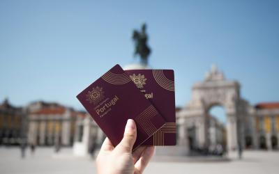 A hand holding two maroon passports with "Portugal" printed on the cover, set against a blurred background of a large plaza with a triumphal arch and a statue on a column.