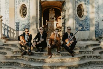 Four musicians posing on stone steps outside a building with blue and white azulejo tiles and circular windows.