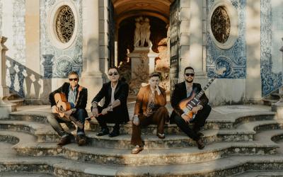 Four musicians posing on stone steps outside a building with blue and white azulejo tiles and circular windows.