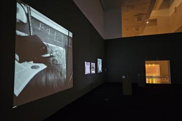 A dimly lit gallery interior at the Centro Cultural de Belém (CCB) in Lisbon, featuring a large projected black-and-white video on the left wall, three smaller framed artworks on the adjacent wall, and an illuminated doorway leading to another roo