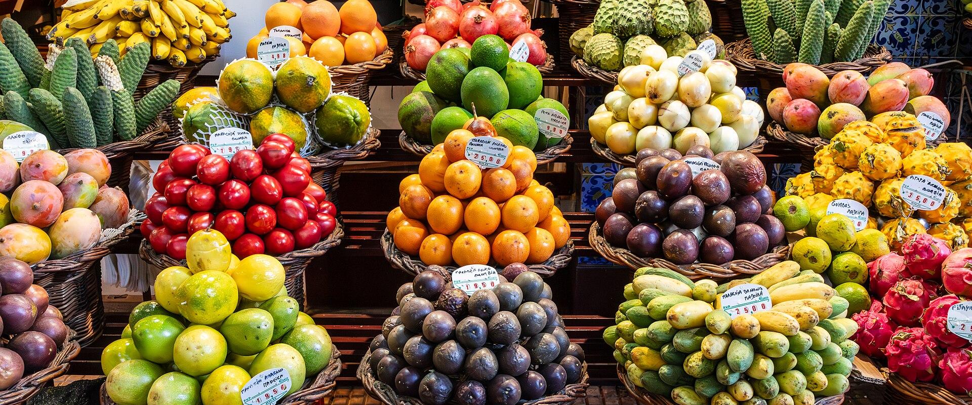 A vibrant display of exotic fruits, including dragon fruit, passion fruit, and various citrus, arranged in woven baskets on wooden shelves, with blue and white patterned tiles visible in the background.