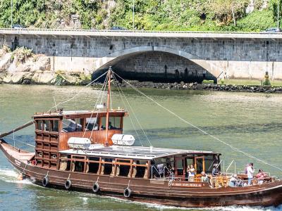 A traditional wooden Rabelo boat carrying tourists cruises on a river with a large stone arch bridge in the background.