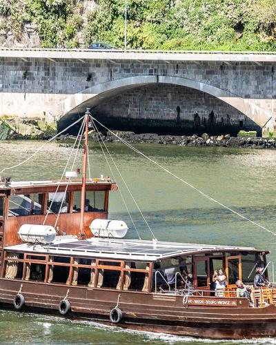 A traditional wooden Rabelo boat carrying tourists cruises on a river with a large stone arch bridge in the background.