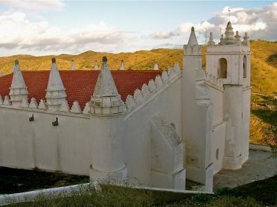 A white-washed church building with a distinctive red-tiled roof, conical buttresses, and a bell tower, set against a backdrop of rolling golden hills.