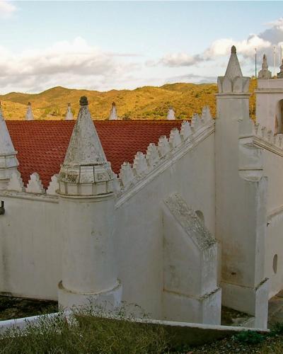 A white-washed church building with a distinctive red-tiled roof, conical buttresses, and a bell tower, set against a backdrop of rolling golden hills.