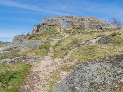 The image shows a stone ruin, specifically a wall with a central arched opening, situated on a rocky, grassy hilltop under a blue sky.