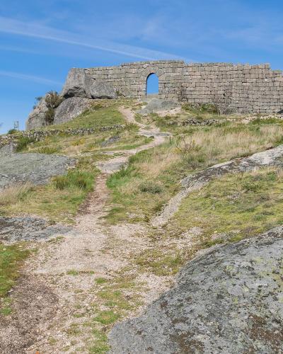 The image shows a stone ruin, specifically a wall with a central arched opening, situated on a rocky, grassy hilltop under a blue sky.