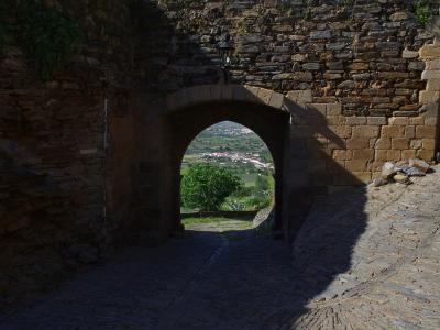 A stone archway in a fortified wall with a cobblestone path leading through it, framing a view of a village and green landscape in the distance.