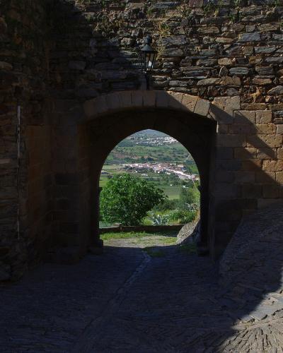 A stone archway in a fortified wall with a cobblestone path leading through it, framing a view of a village and green landscape in the distance.