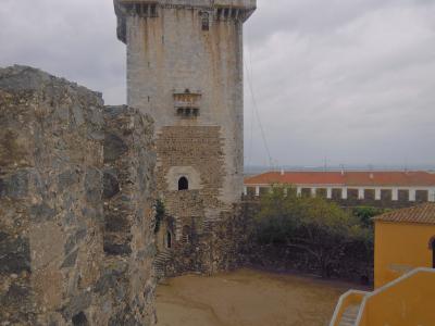 A tall, rectangular stone keep with crenellated battlements and a small balcony, situated within a castle courtyard featuring stone walls and a yellow building.