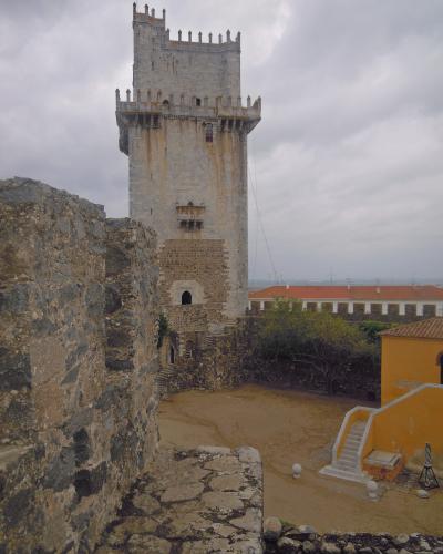 A tall, rectangular stone keep with crenellated battlements and a small balcony, situated within a castle courtyard featuring stone walls and a yellow building.