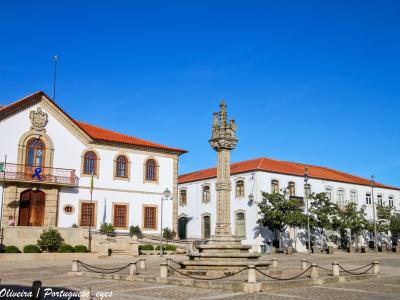 The image shows a sunlit town square in Portugal featuring a prominent stone pillory (pelourinho) in the center, flanked by white buildings with red-tiled roofs, including a municipal building with a Portuguese flag and a blue ribbon; the photo is